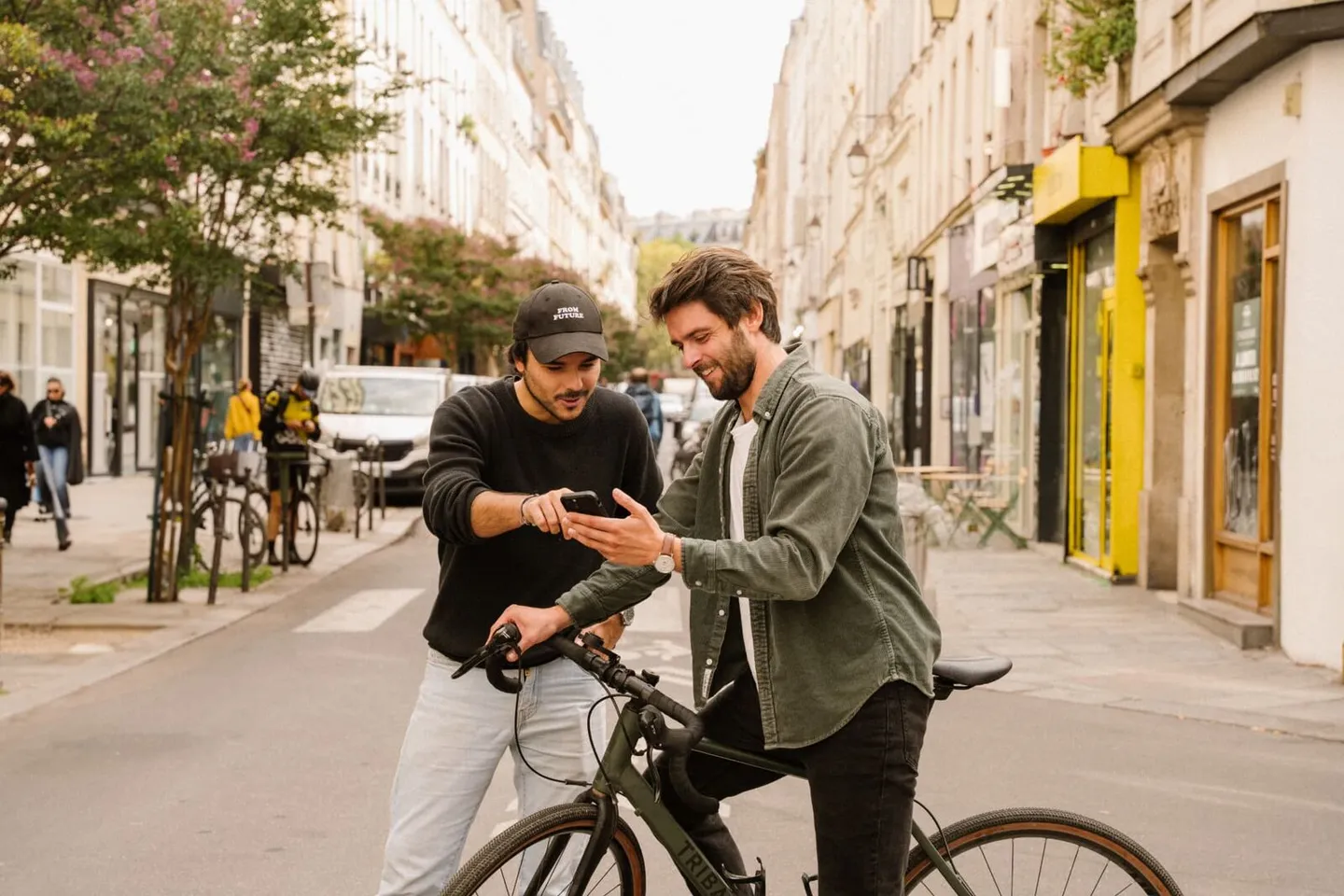 Deux amis dans une rue, l'un sur son vélo, regardent ensemble un téléphone